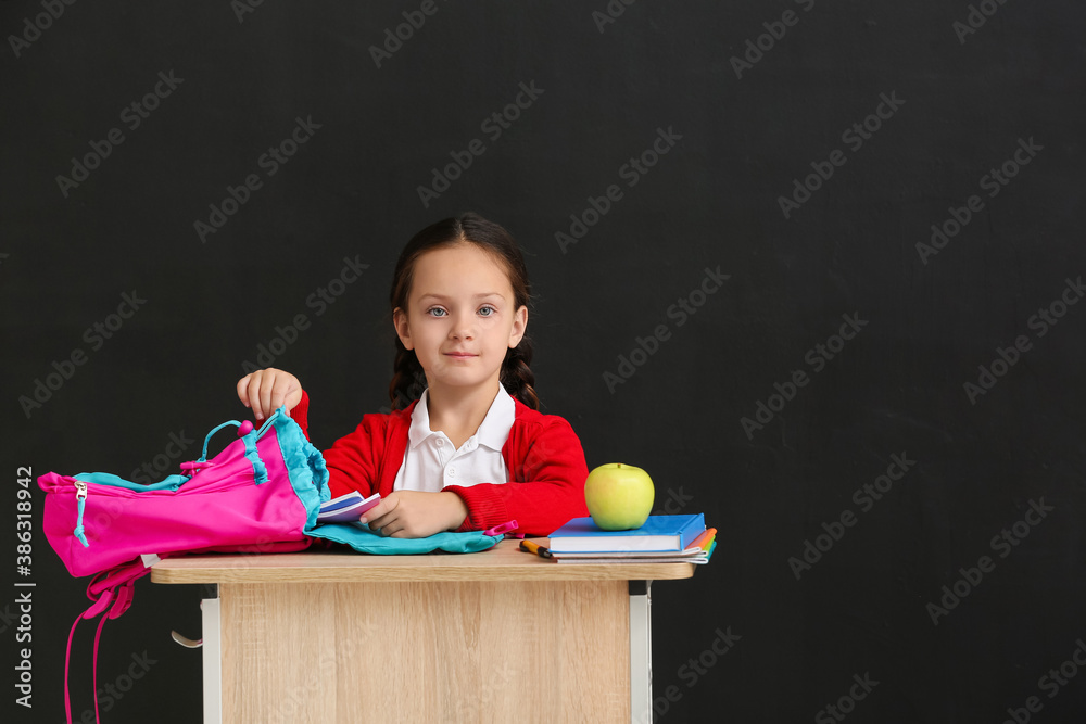 Little pupil sitting at school desk against dark background