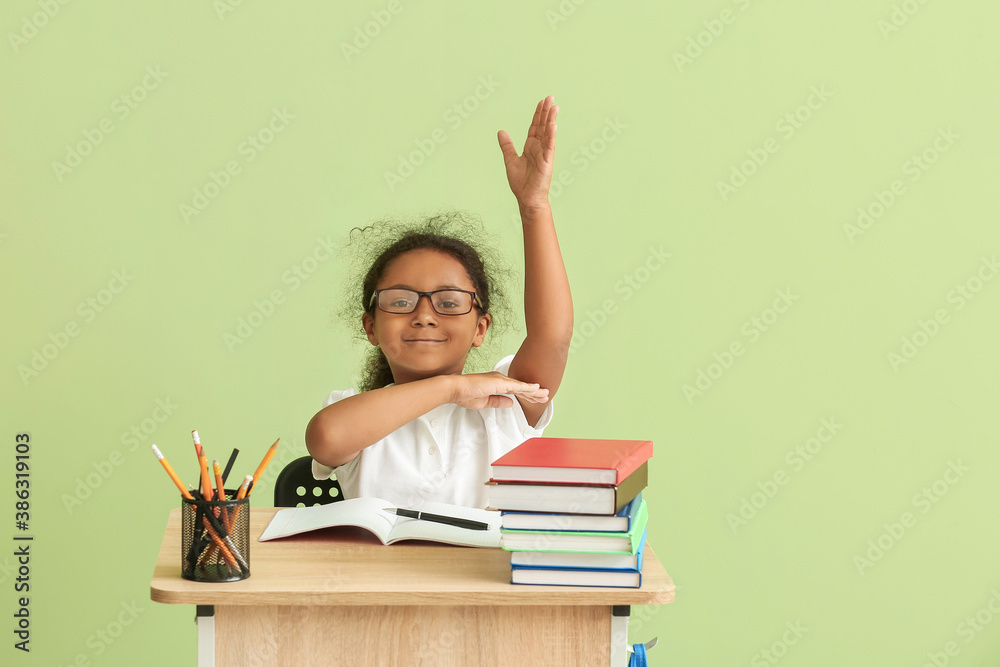 Cute African-American schoolgirl with raised hand sitting at desk against color background