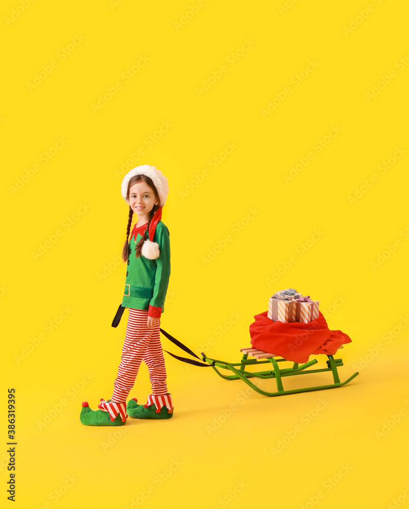 Cute little girl dressed as elf, with sledge and Christmas gifts on color background