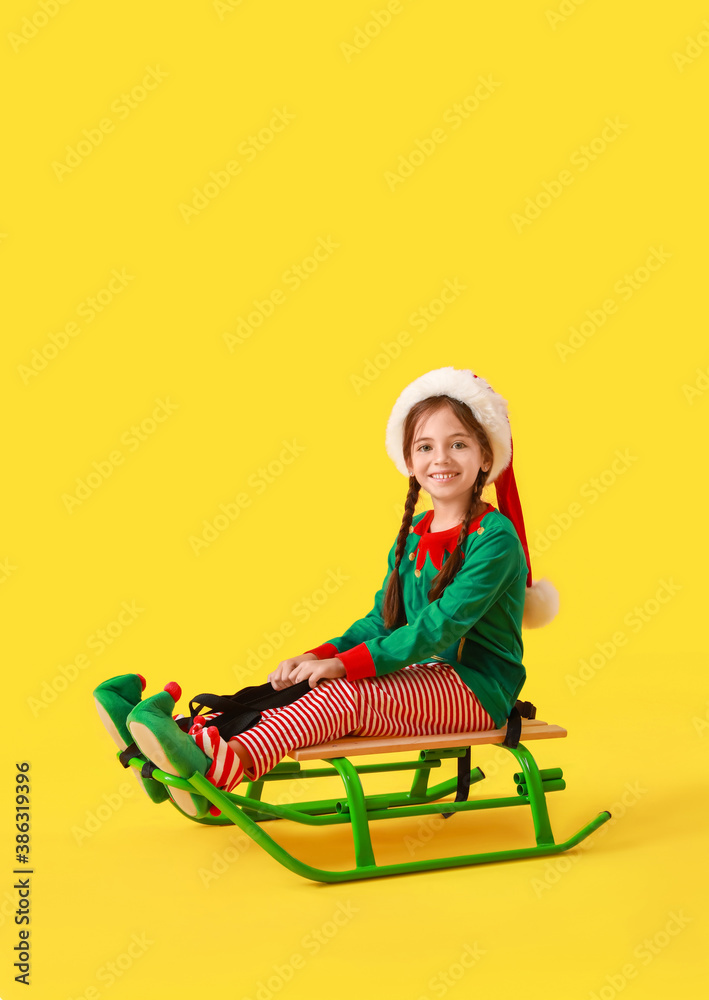 Cute little girl dressed as elf sitting on sledge against color background