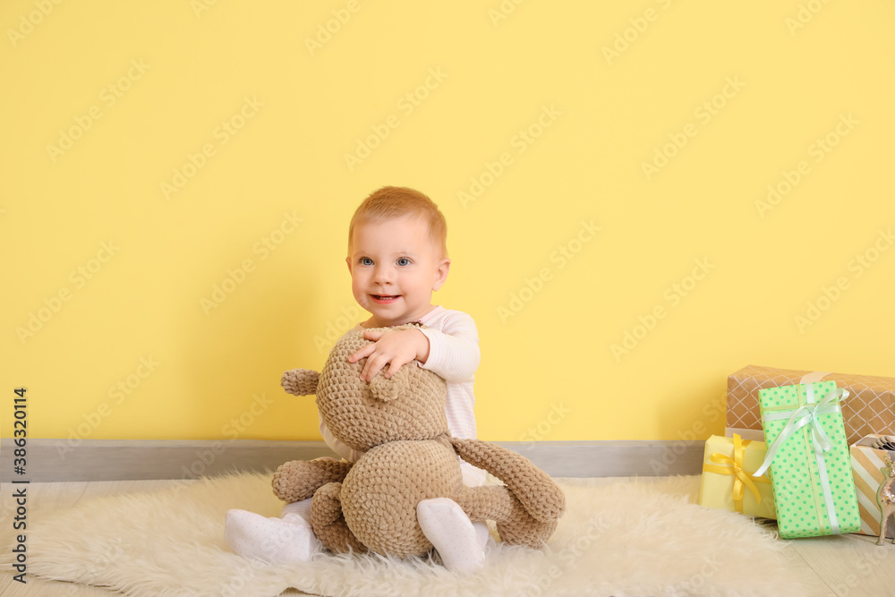 Cute little baby with toy and Christmas gifts near color wall