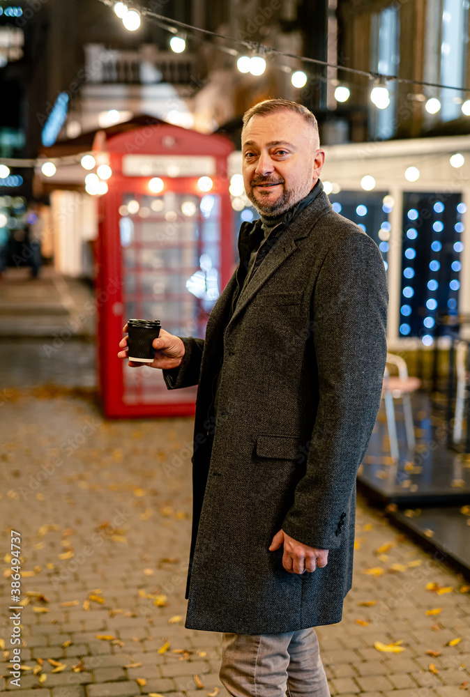 Coffee break. Mature man in elegant coat smiling nicely while having coffee. Evening street.