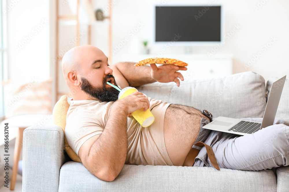 Overweight man with laptop eating unhealthy food while lying on sofa