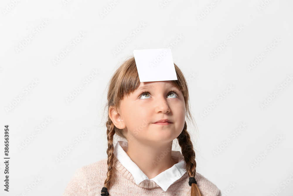 Little girl with blank note paper on her forehead against light background