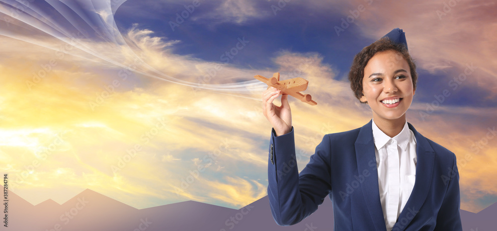 Beautiful African-American stewardess holding wooden airplane against blue cloudy sky