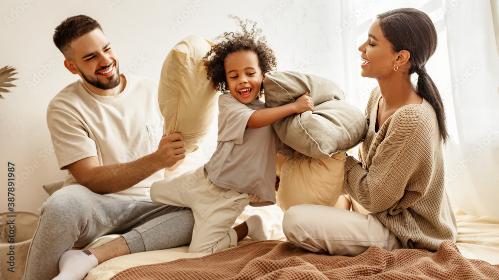 happy multiethnic family mom, dad and child  laughing, playing, fights pillows and jumping in bed   