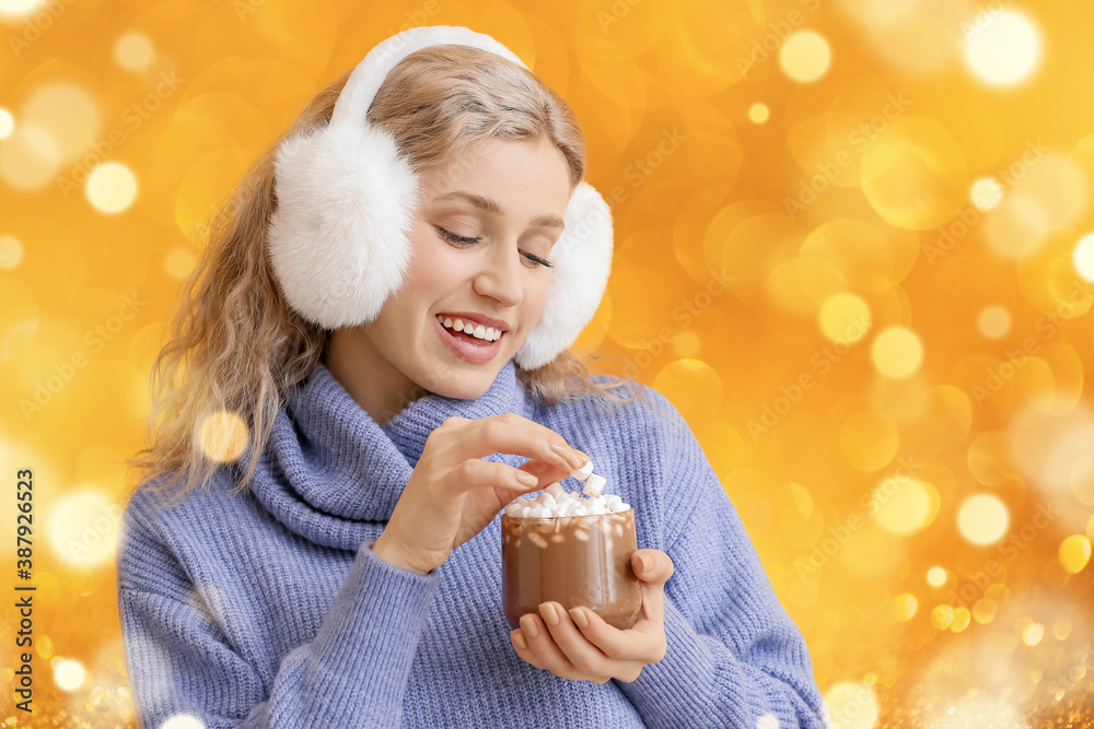 Beautiful young woman with fur headphones and cup of hot chocolate on color background