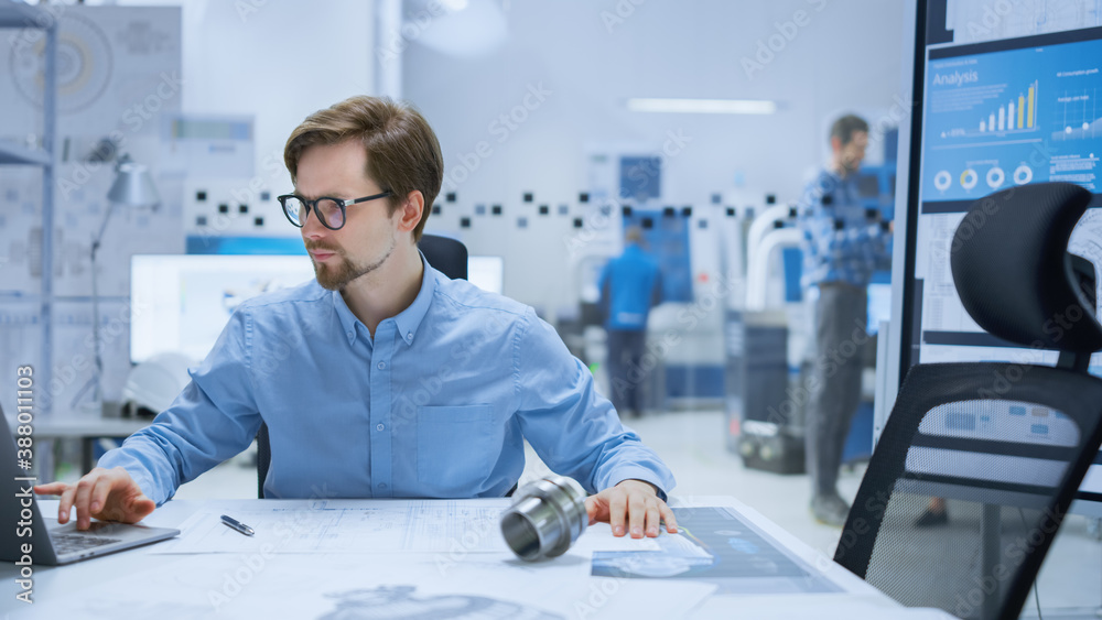 Modern Factory: Industrial Engineer Sitting at His Desk in Workshop, Working on Computer, Redrawing 