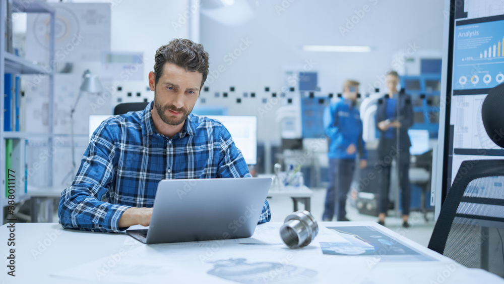 
Modern Industrial Factory: Industrial Engineer Sitting at His Desk, Working on Laptop Computer, Ana