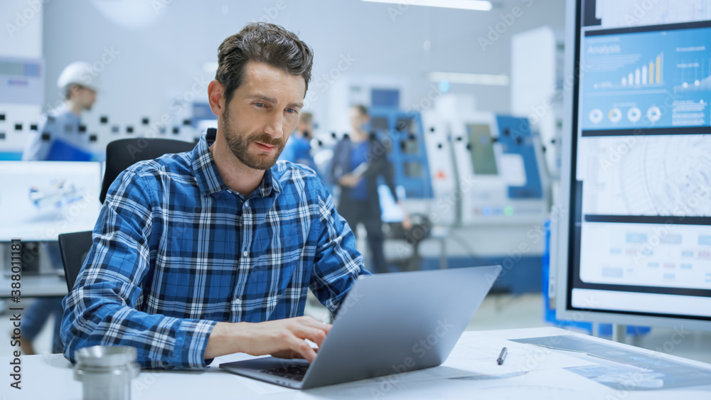 Modern Industrial Factory: Portrait of Industry Engineer Sitting at His Desk, Working on Laptop, Ana