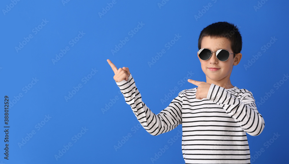 Cute boy with stylish sunglasses pointing at something against color background