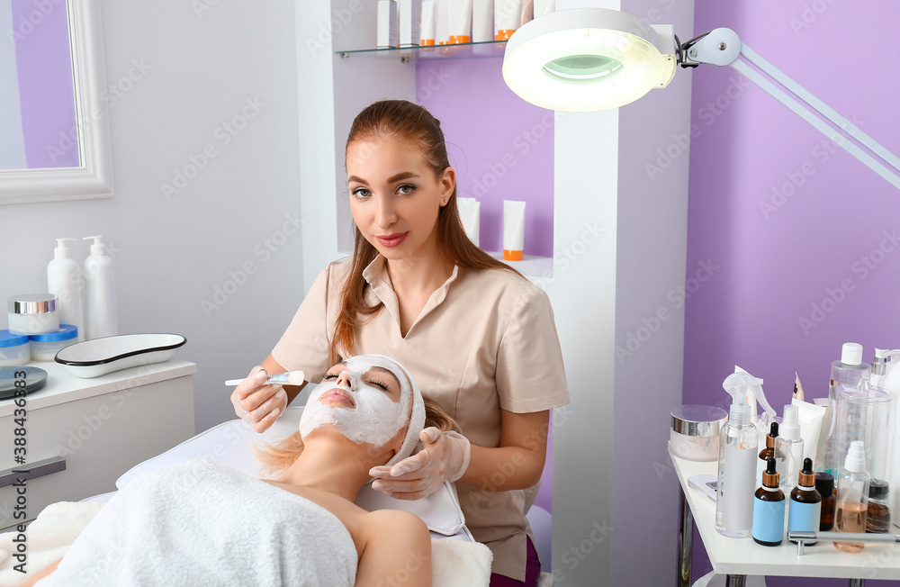 Cosmetologist applying mask onto face of woman in beauty salon