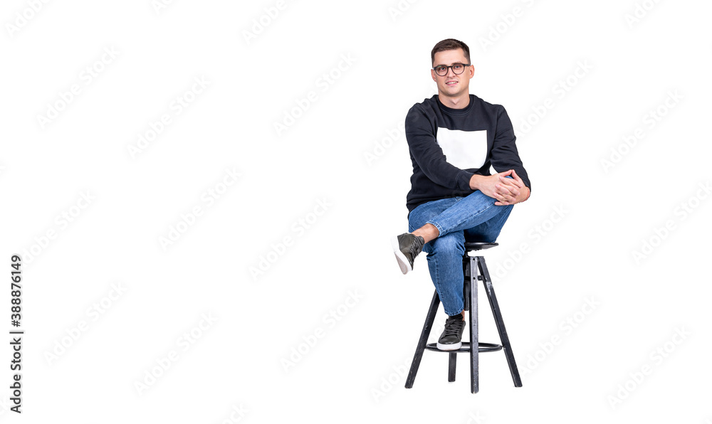 A young attractive guy in blue and black clothes sits on a bar stool on a white background and looks