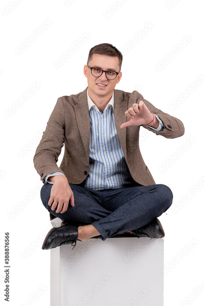 Young man in suit sitting on the floor with crossed hands. Showing thumb down. White background.