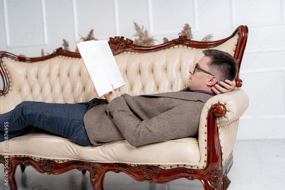 Portrait of a handsome fashionable man posing in the interior. Male resting on sofa with books in ha