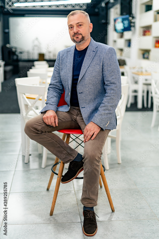 Seriuos bearded man in suit sits on bar chair. Grey jacket and light brown pants. Modern interior ba