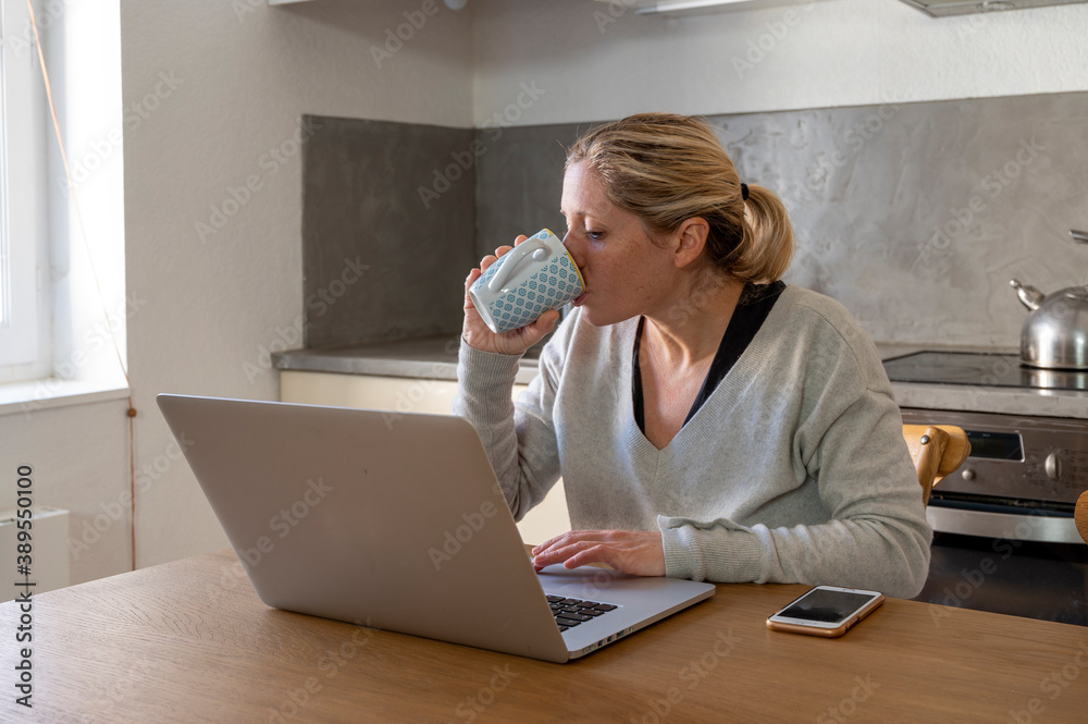 Femme dans sa cuisine en télétravail devant son ordinateur buvant un café