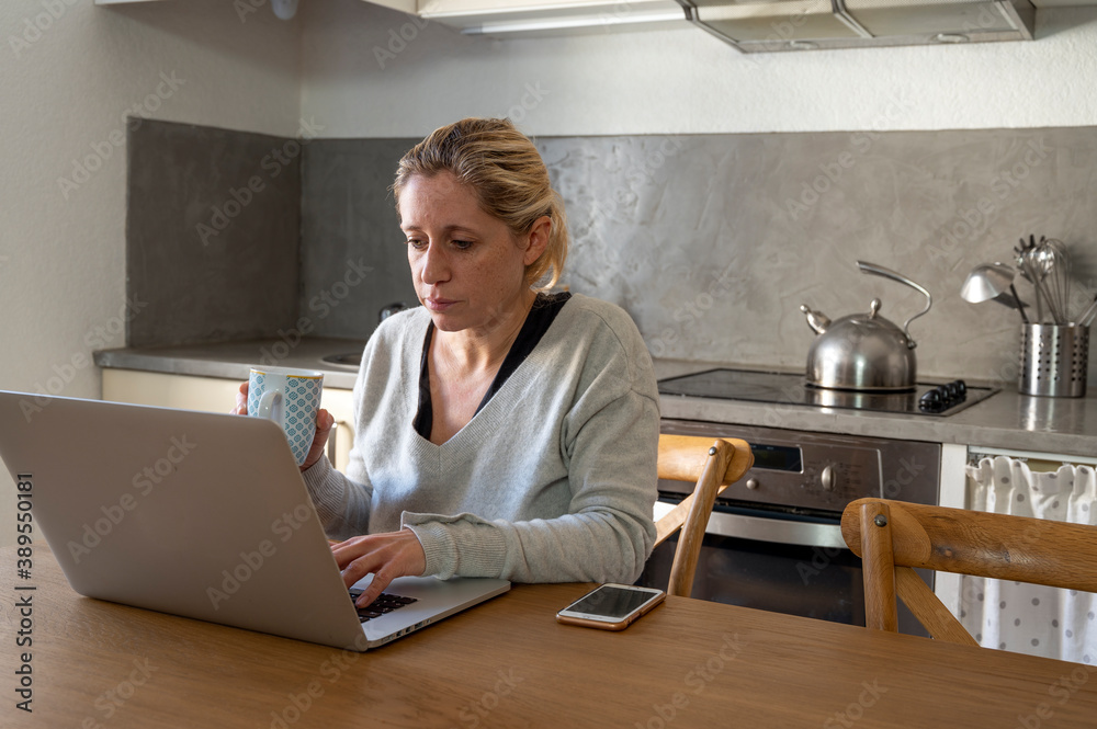 Femme dans sa cuisine en télétravail devant son ordinateur buvant un café