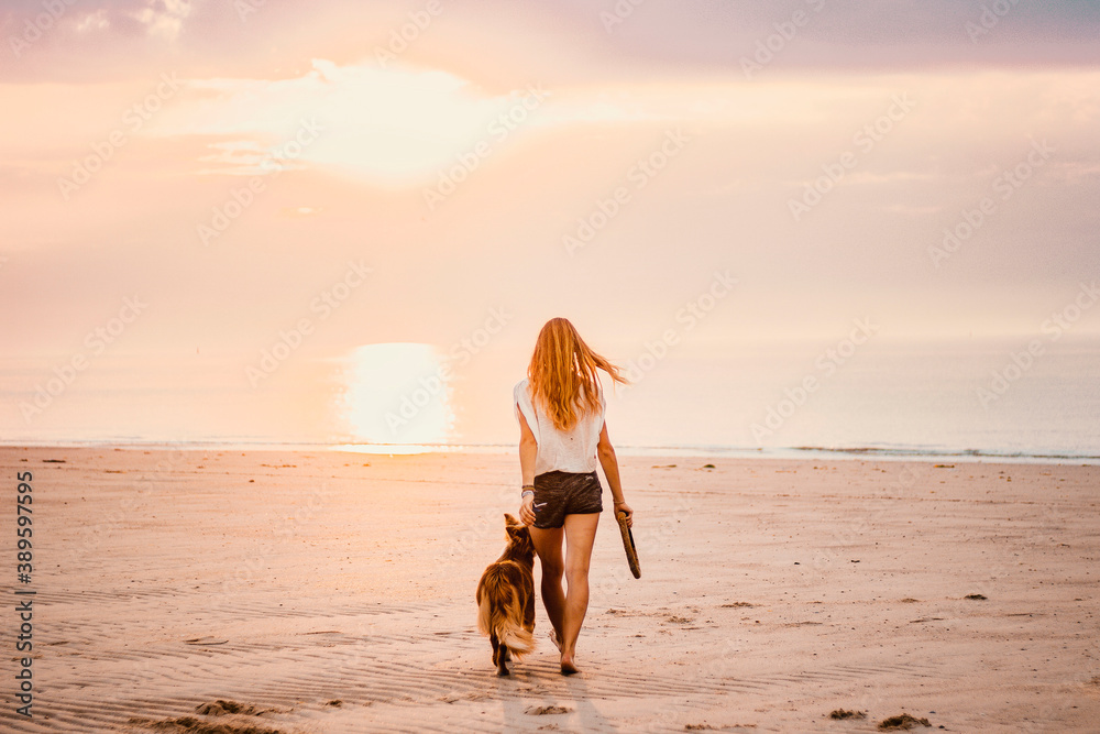 Young blond woman walking with her pet border collie dog on a golden sandy beach during a sunset