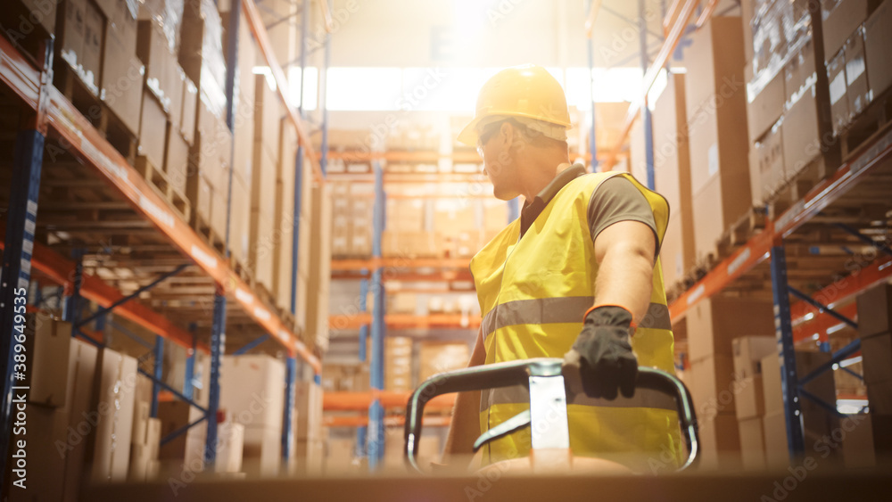 Worker Moves Cardboard Boxes using Hand Pallet Truck, Walking between Rows of Shelves with Goods in 