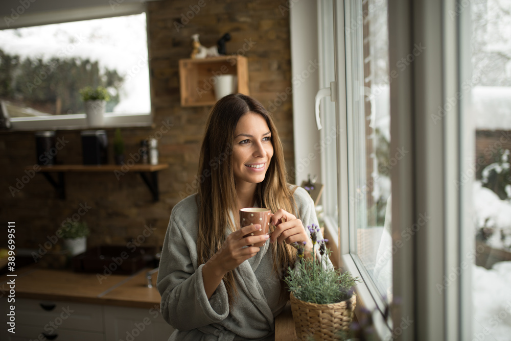 Gorgeous young woman standing in the kitchen drinking coffee and looking through window on winter da