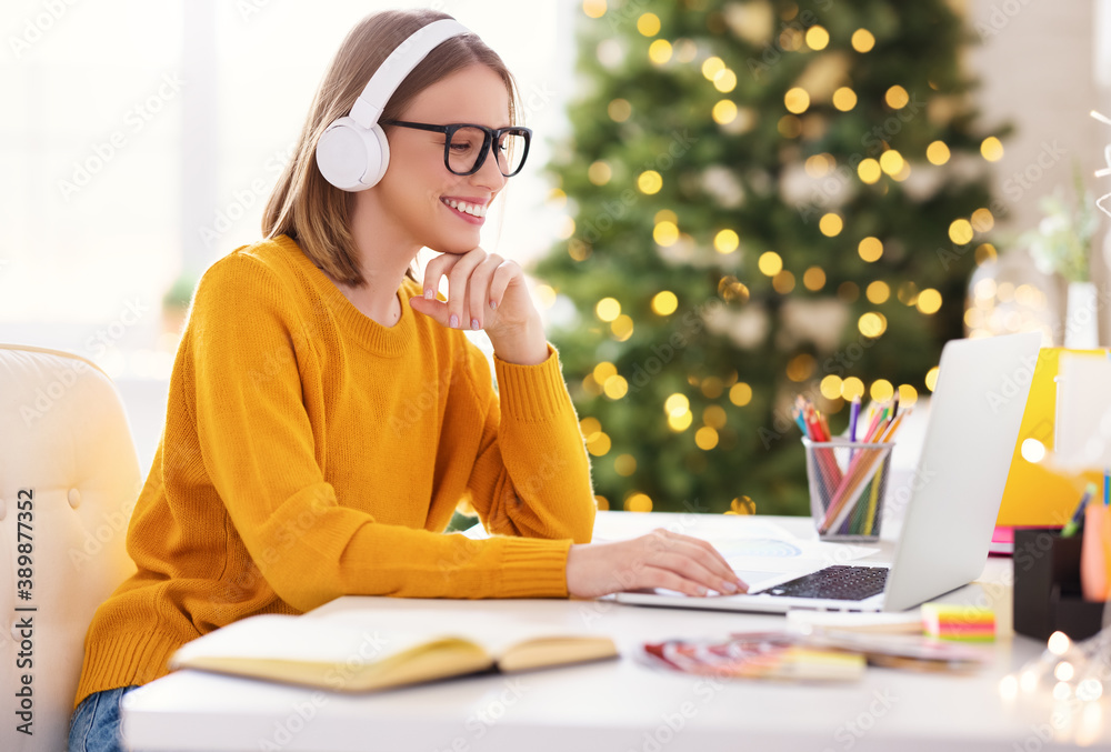 Happy young woman freelancer in headphones working remote at laptop at home before Christmas.