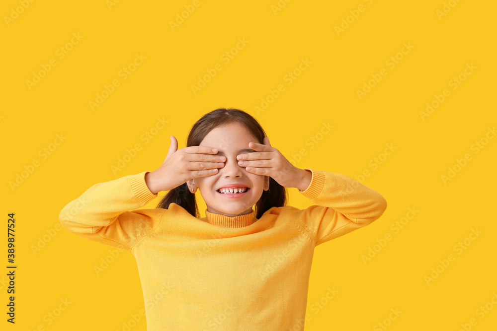 Cute little girl in autumn clothes covering her eyes with hands on color background