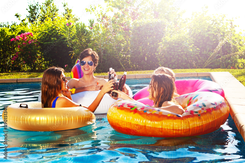 Happy boys and girls swim in the outdoor swimming pool with soda bottles chat partying