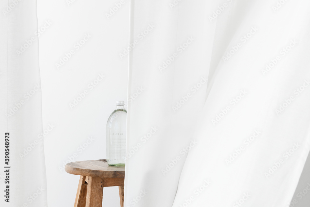 Glass water bottle on a wooden stool behind a flowing white curtain