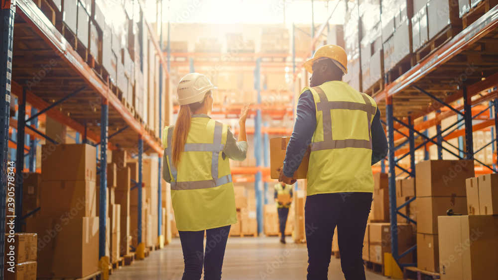 Male and Female Workers Wearing Hard Hats Walking Through Retail Warehouse full of Shelves with Good