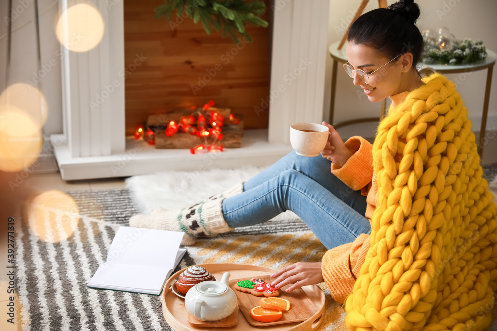 Beautiful young woman with warm plaid reading book and drinking tea at home