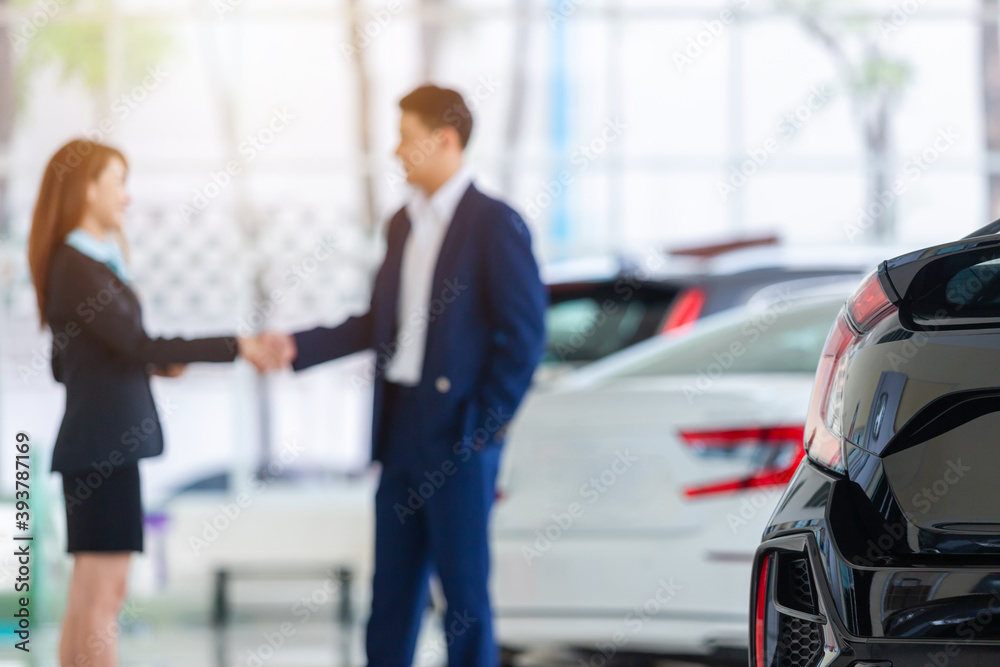 Selective focus on a new car and blur the dealership professional salesman and his client shaking ha