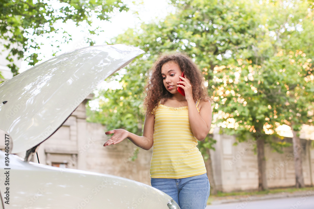 Stressed African-American woman talking by phone near broken car outdoors