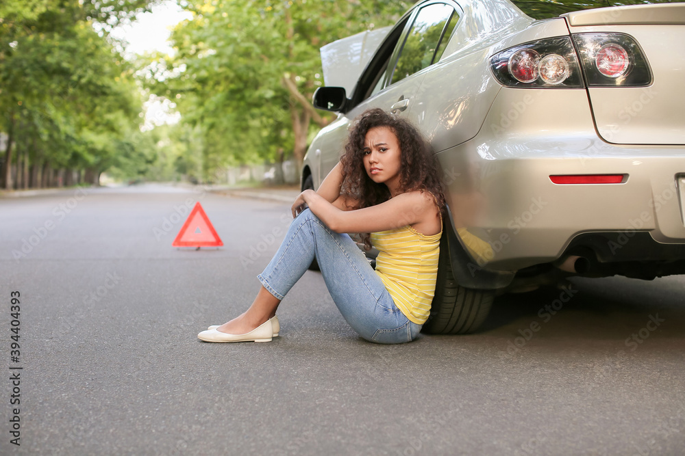 Sad African-American woman sitting near broken car outdoors