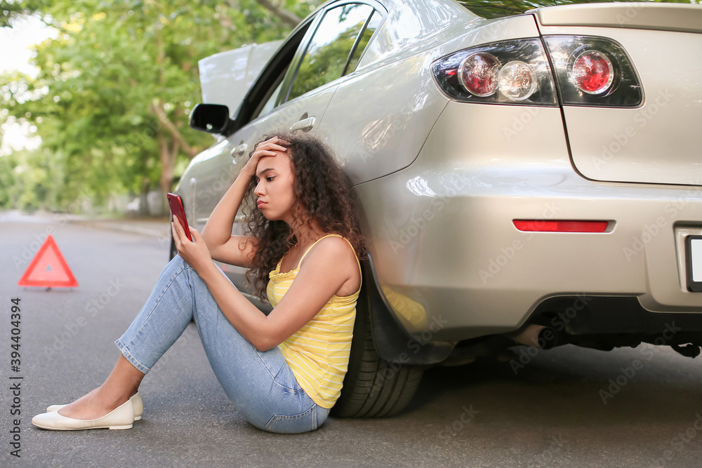 Stressed African-American woman with mobile phone sitting near broken car outdoors