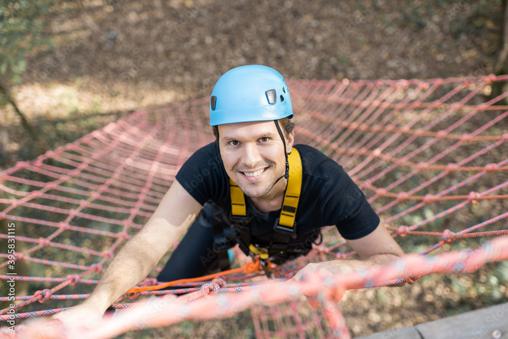 Well-equipped man climbing on a net outdoors, having active recreation in amusement park