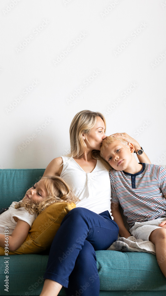 Blonde mom kissing her son’s head and relaxing with daughter on the couch blank space