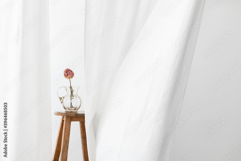 Dry peony flower in a glass jug on a wooden stool in a white room