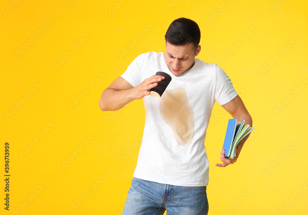 Stressed young man with coffee stains on his t-shirt holding notebooks on color background