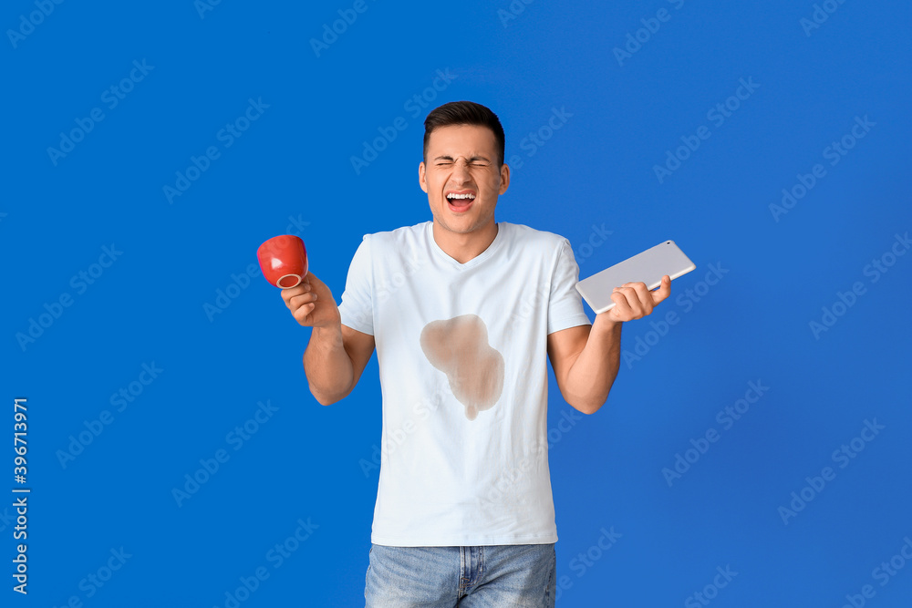 Screaming young man with coffee stains on his t-shirt holding tablet on color background