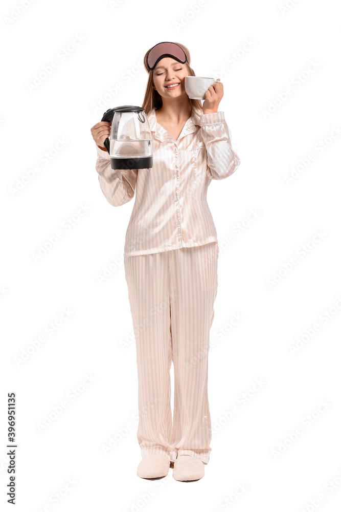 Young woman in pajamas holding electric kettle and cup on white background