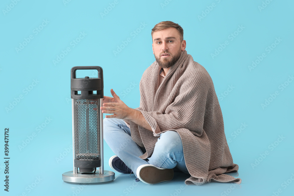 Young man warming hands near electric heater on color background. Concept of heating season