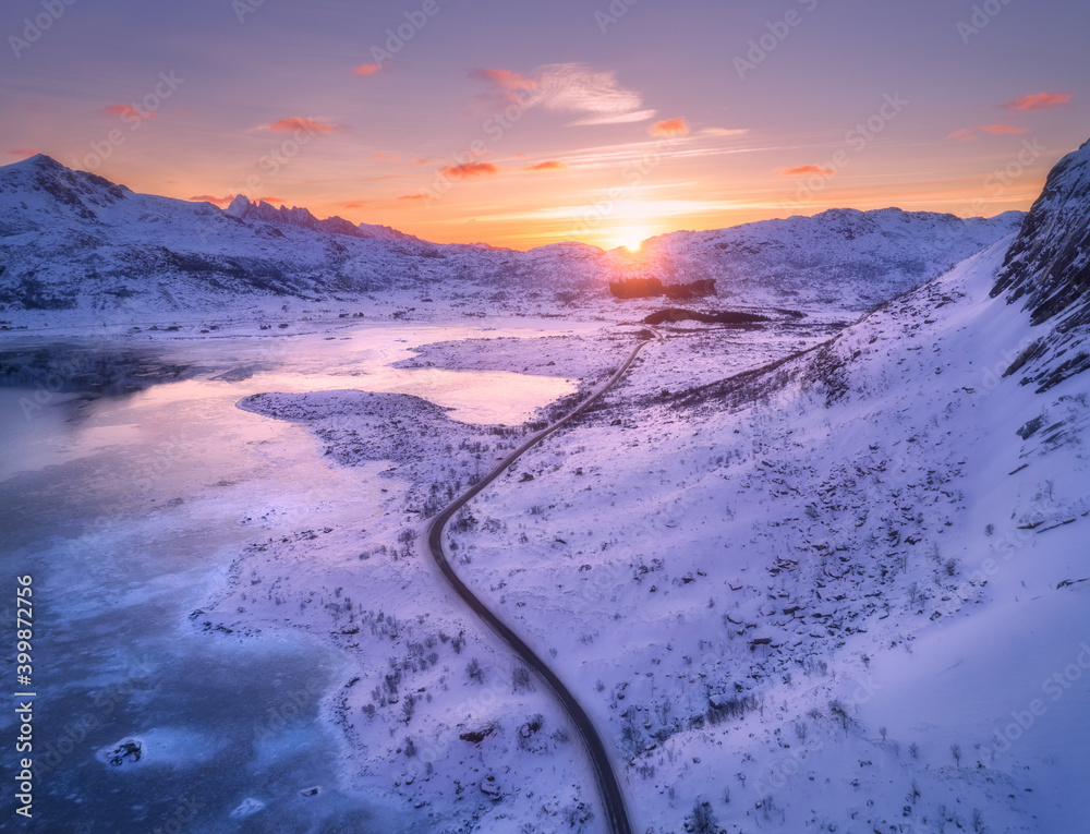 鸟瞰蜿蜒的道路，白雪覆盖的山脉，冬季日落时的橙色天空。景观与