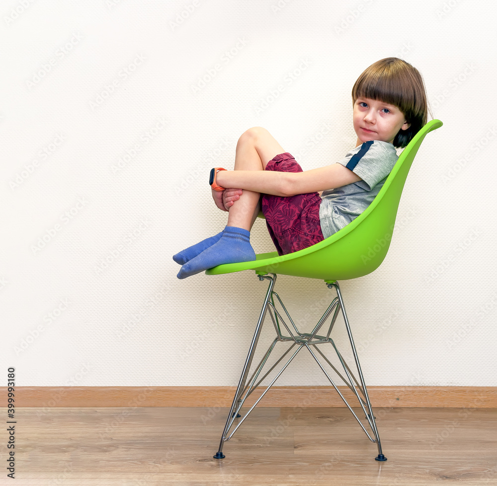 A boy is sitting on a green сhair in profile a front of the white wall. Side view.