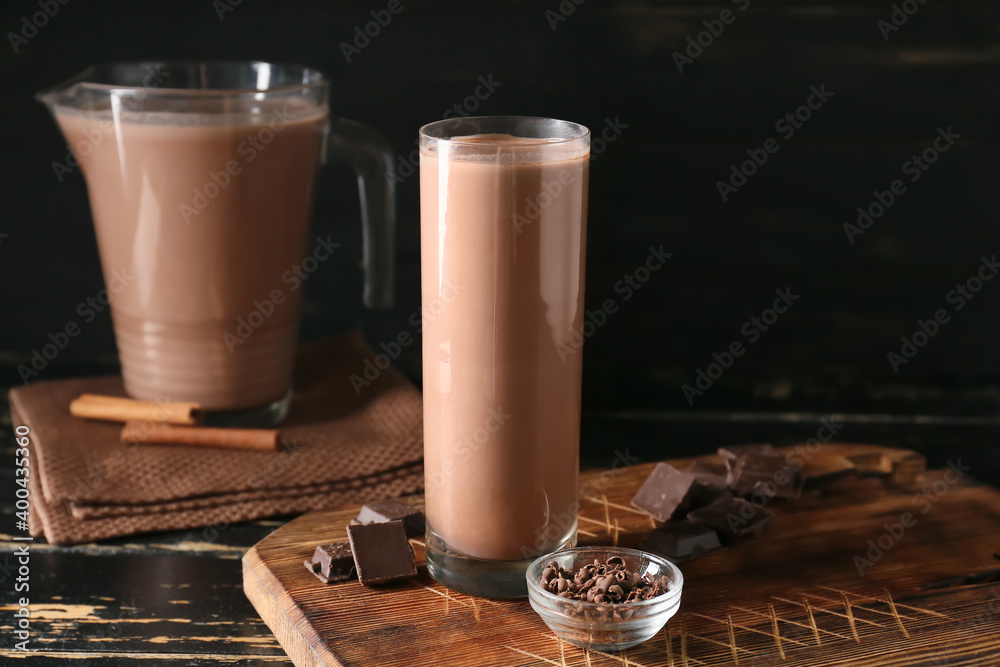 Wooden board with glass of tasty chocolate milk on dark background