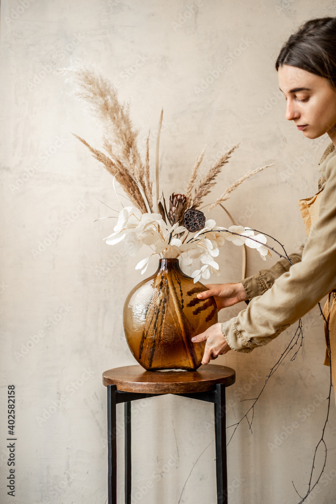 Woman decorating home with a composition of dried flowers and herbs in a glass vase on a beige wall 