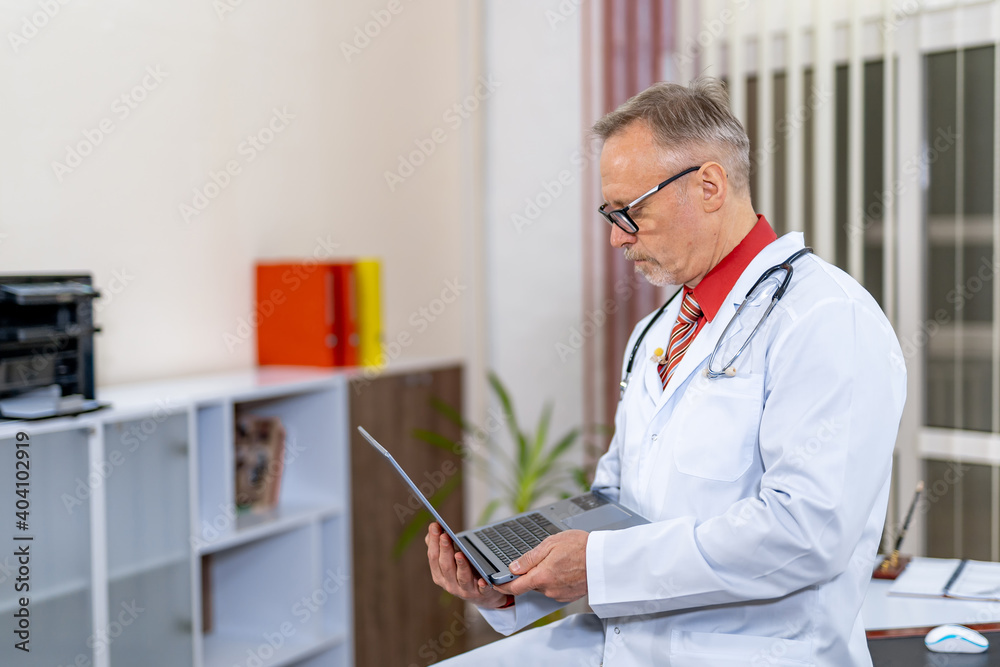 Doctor sits on table end with laptop in hands. Medic types prescription in clinic office.