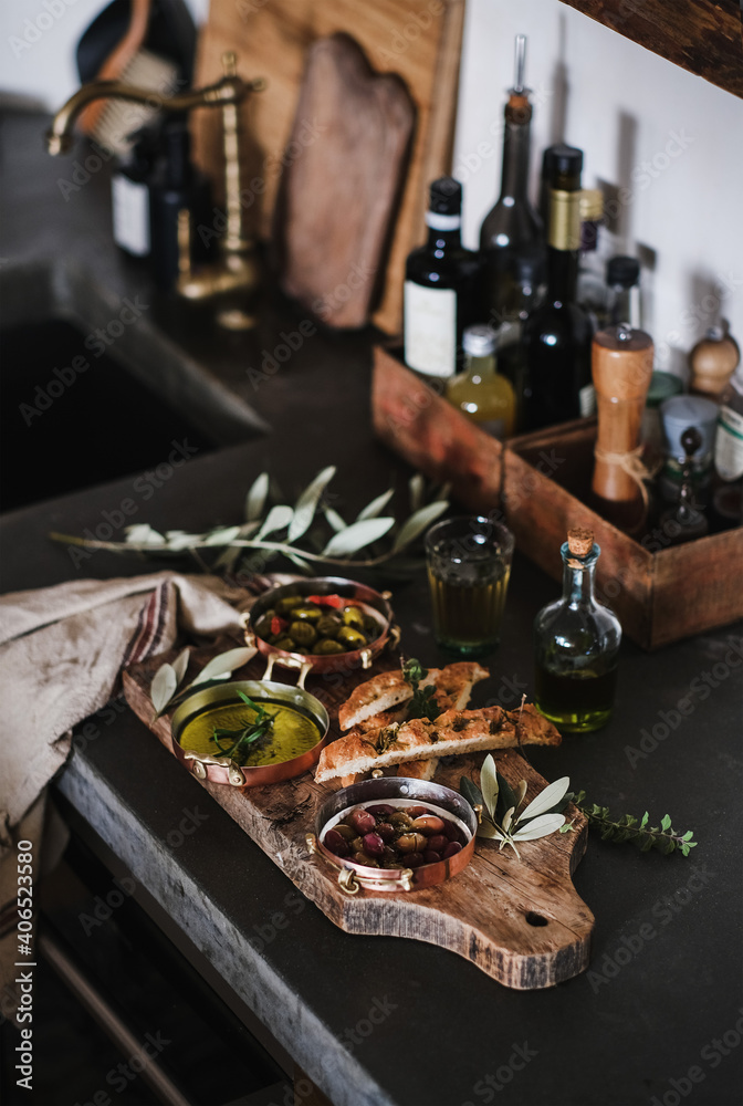 Pickled Greek olives, olive oil in copper jars and herbed focaccia slices on rustic wooden board ove