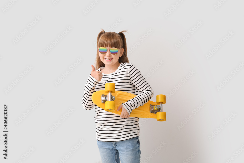 Cute little girl with stylish sunglasses and skateboard showing thumb-up gesture on light background