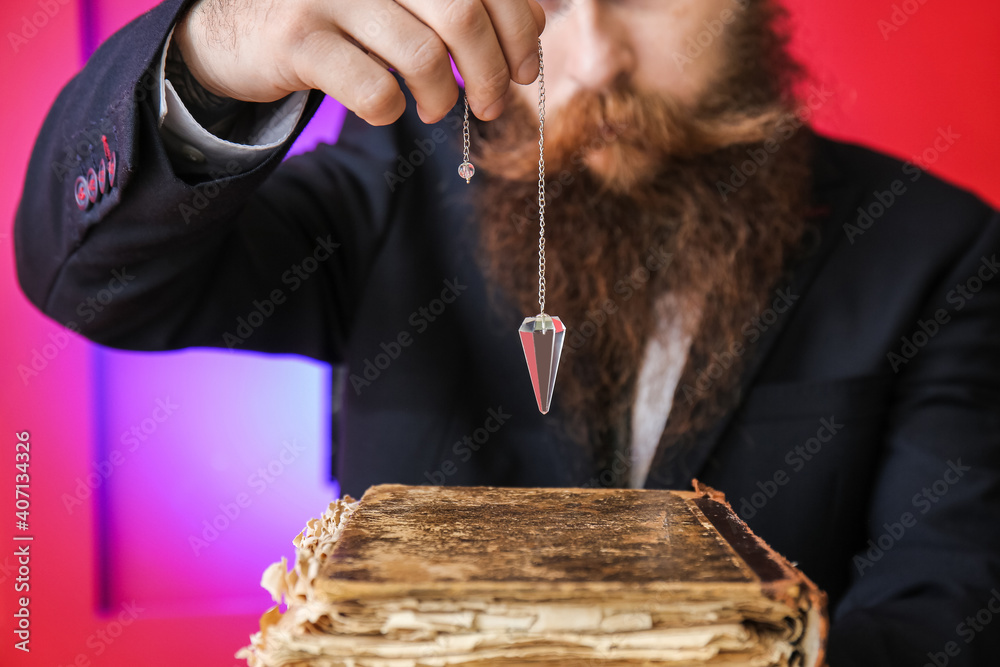 Male fortune teller with book and pendulum on color background